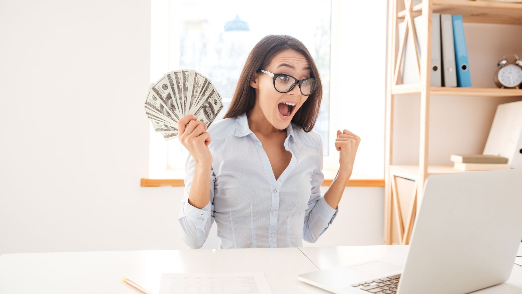 Young woman smiling while holding cash and using a laptop